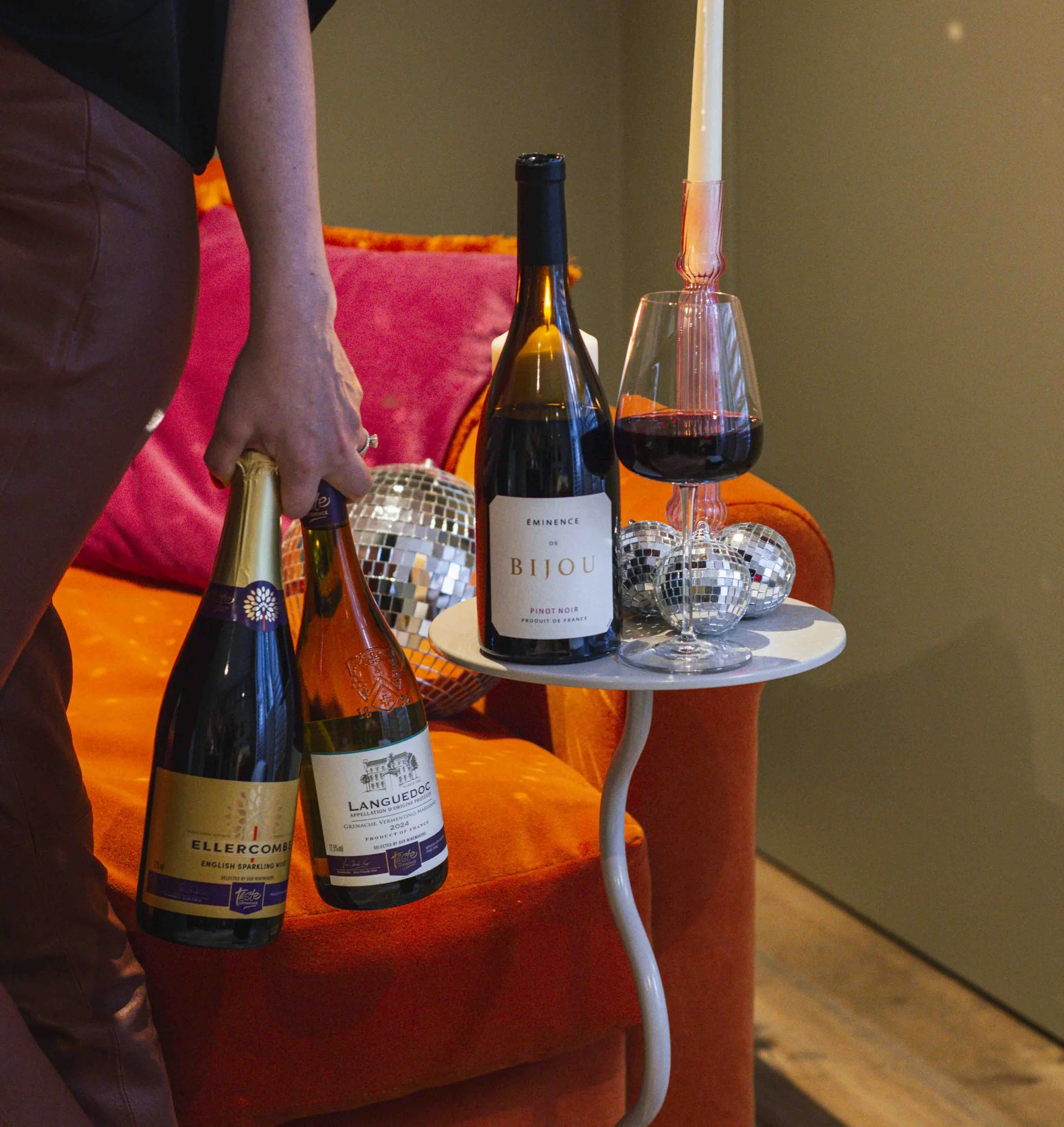 Wine bottles and a red wine glass beside a lit candle in a colourful lounge setting