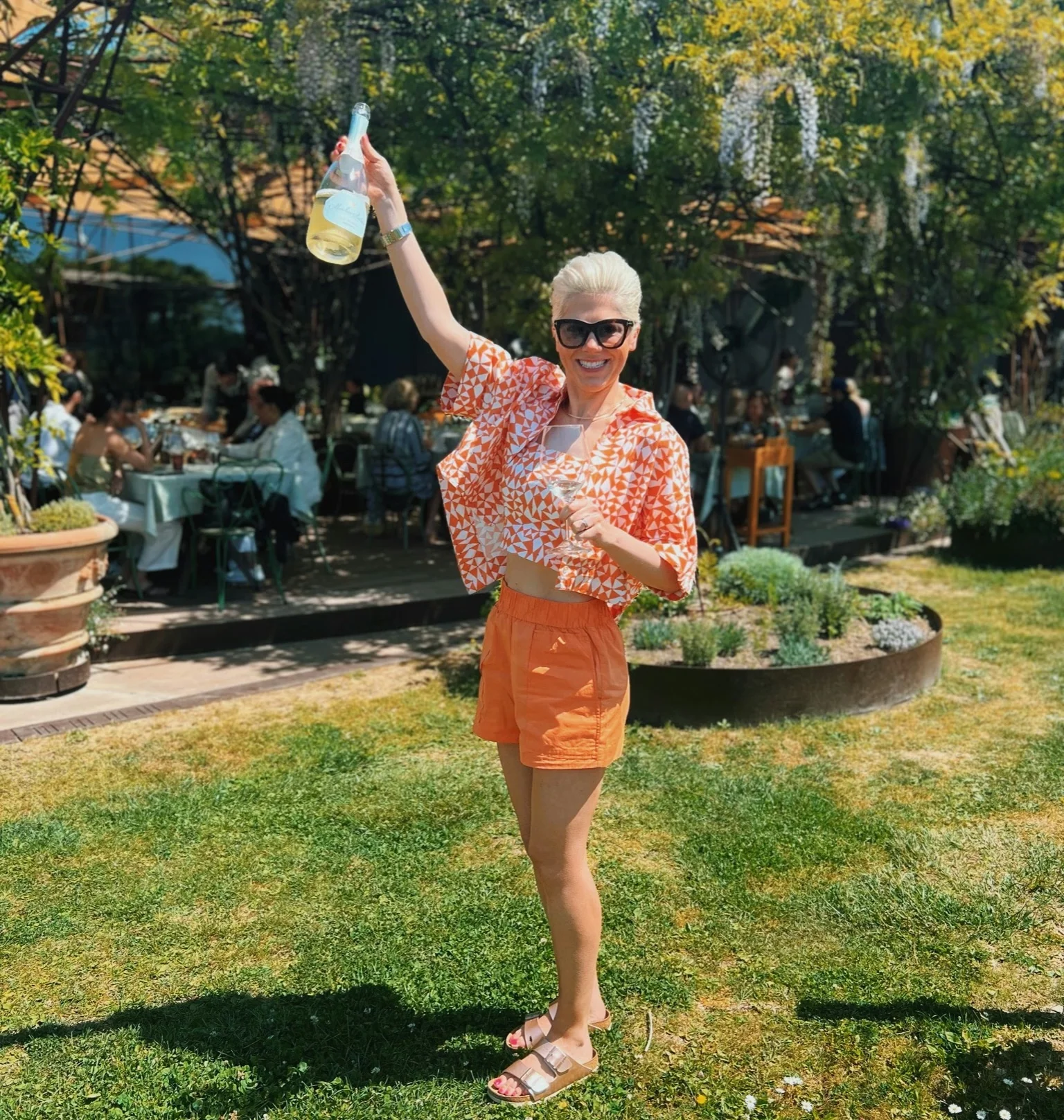 Woman raising a wine bottle in a sunny Tuscan winery garden, with outdoor tables and trees behind her