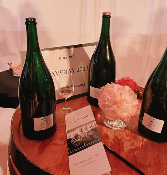 Three Bollinger Champagne bottles and a glass on a barrel table with flowers and event leaflets