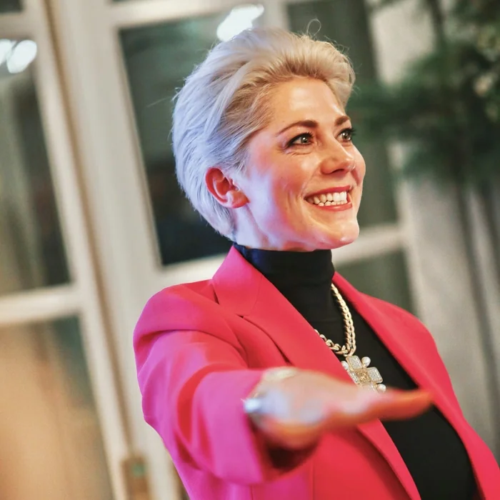 Smiling woman in a bright pink blazer gestures towards the camera indoors.