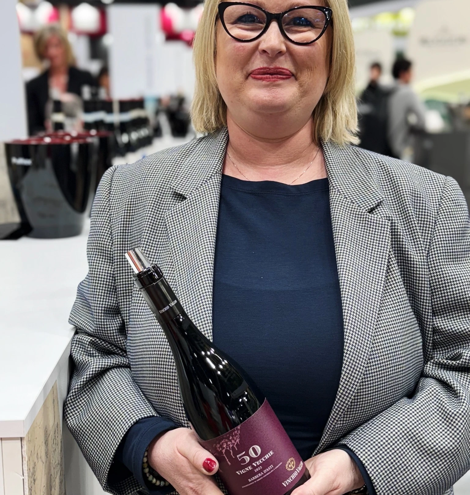 Elizabeth holds a bottle of 50 Vigne Vecchie wine at Wine Paris, with the exhibition hall behind her