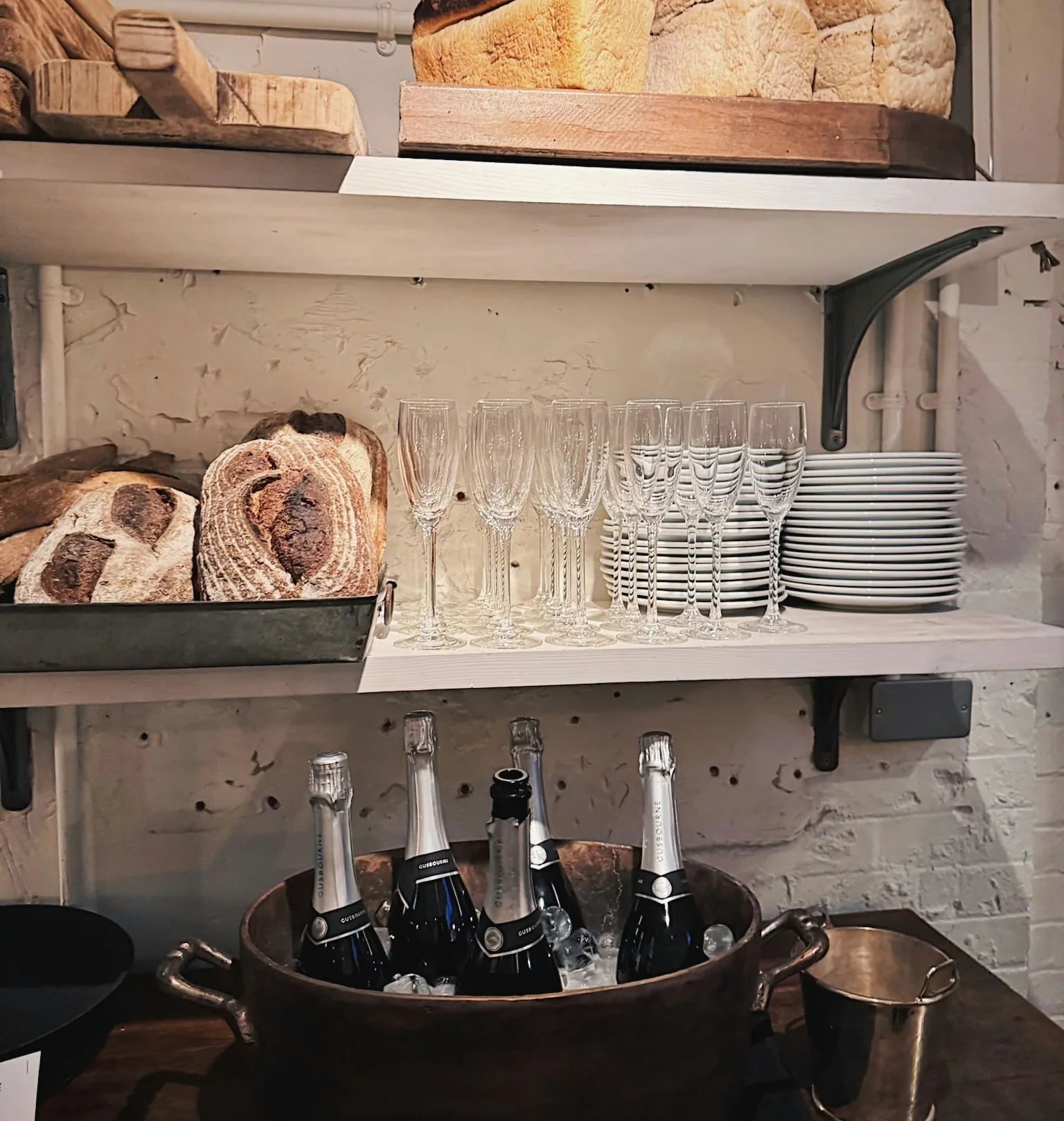 Chilled sparkling wine bottles beside bread, champagne flutes and stacked plates on wooden shelves