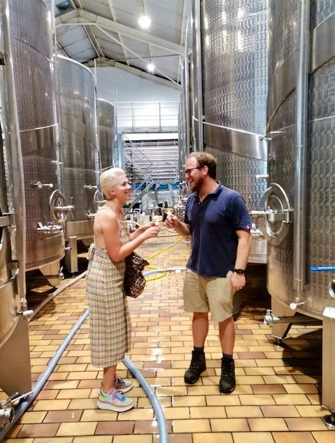 Two people toasting wine in a winery between stainless steel tanks