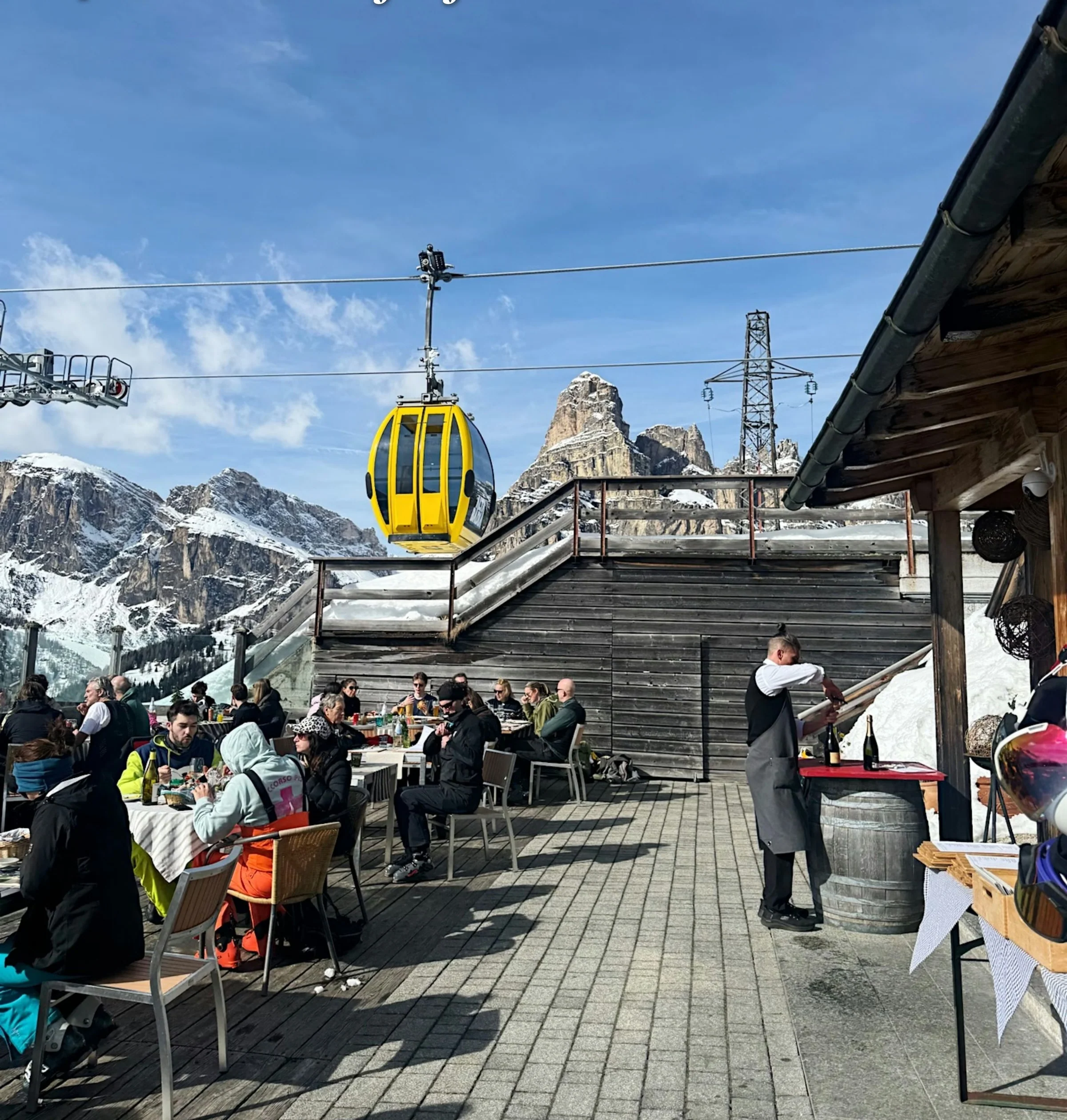 Outdoor mountain restaurant terrace with diners, a yellow cable car and snow-covered Dolomite peaks