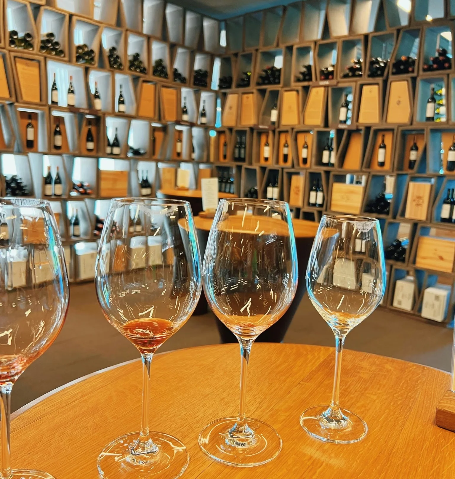 Four wine glasses on a wooden table, with wine bottles displayed on shelves behind them at an Italian winery