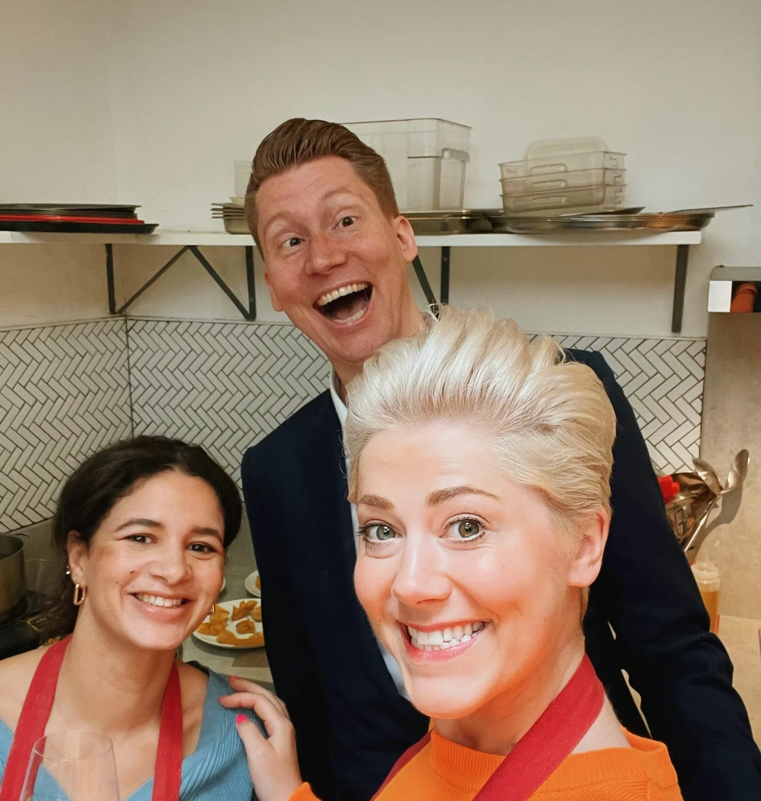 Three smiling people pose together in a kitchen, with a glass of white wine in the foreground
