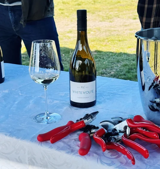 Whitewolfe Chardonnay bottle, wine glass and red-handled corkscrews on a table beside a vineyard