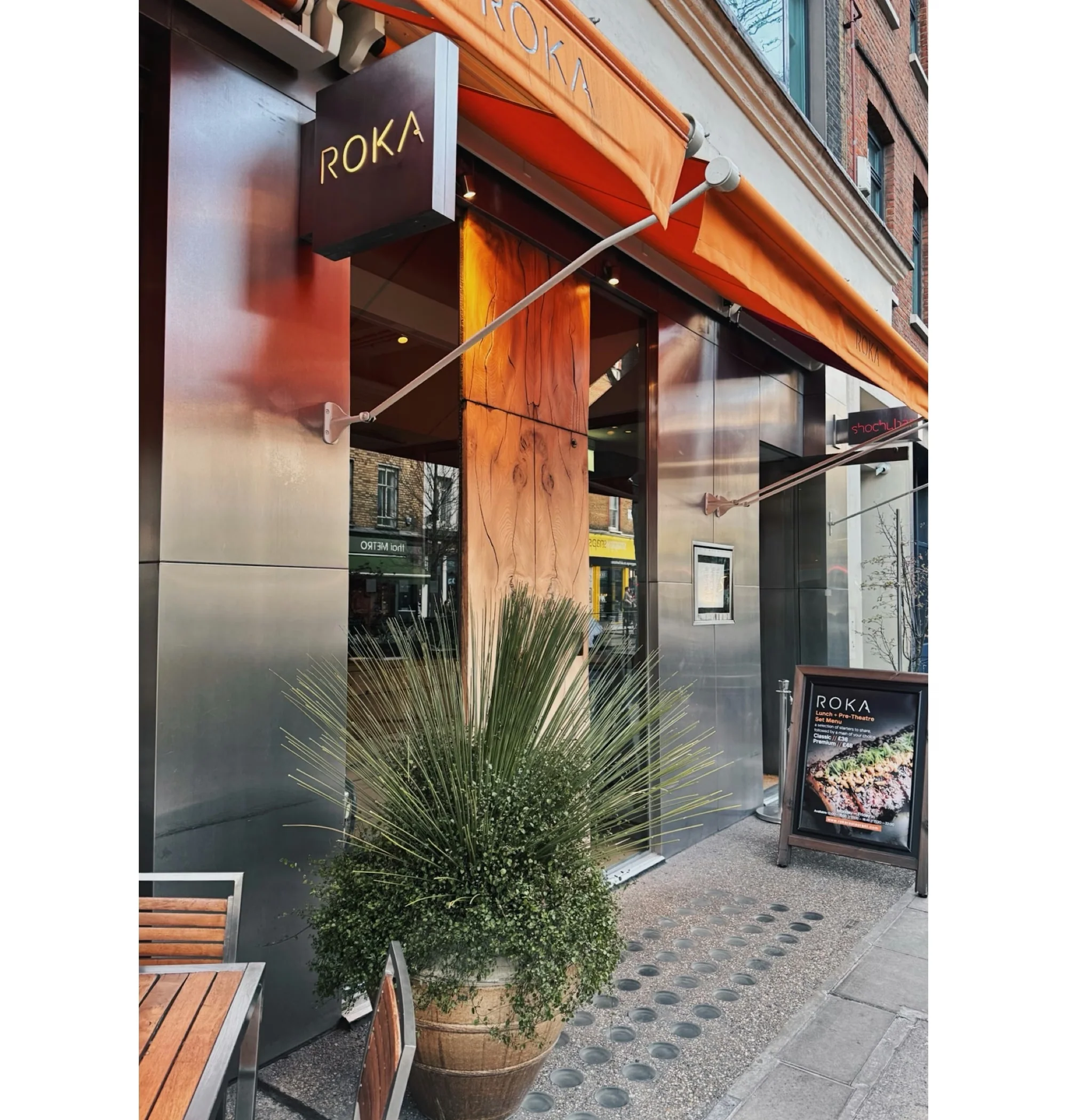 Roka restaurant entrance with orange awning, outdoor seating and a large potted plant