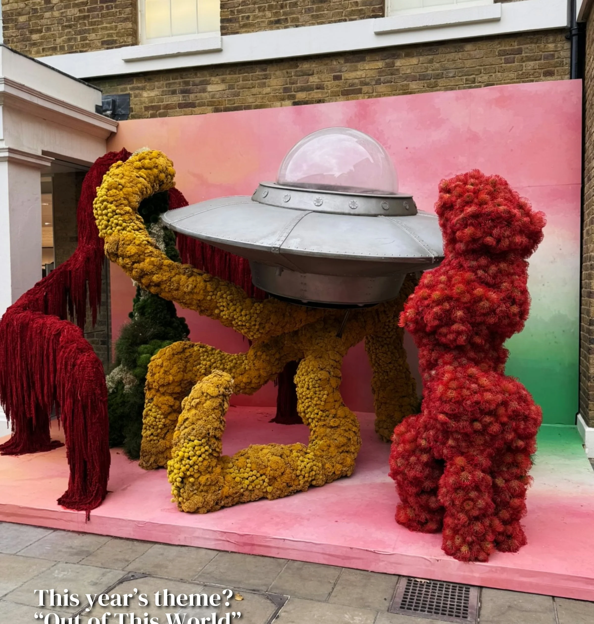 Floral astronaut sculpture displayed against a pink backdrop outside a brick building