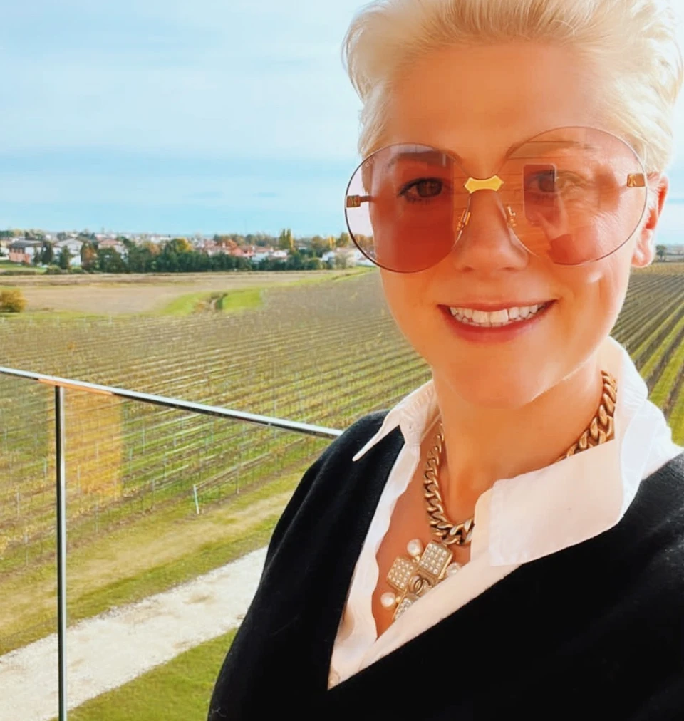 Elizabeth Hawthornthwaite smiling in sunglasses, with vineyard rows and a village behind her