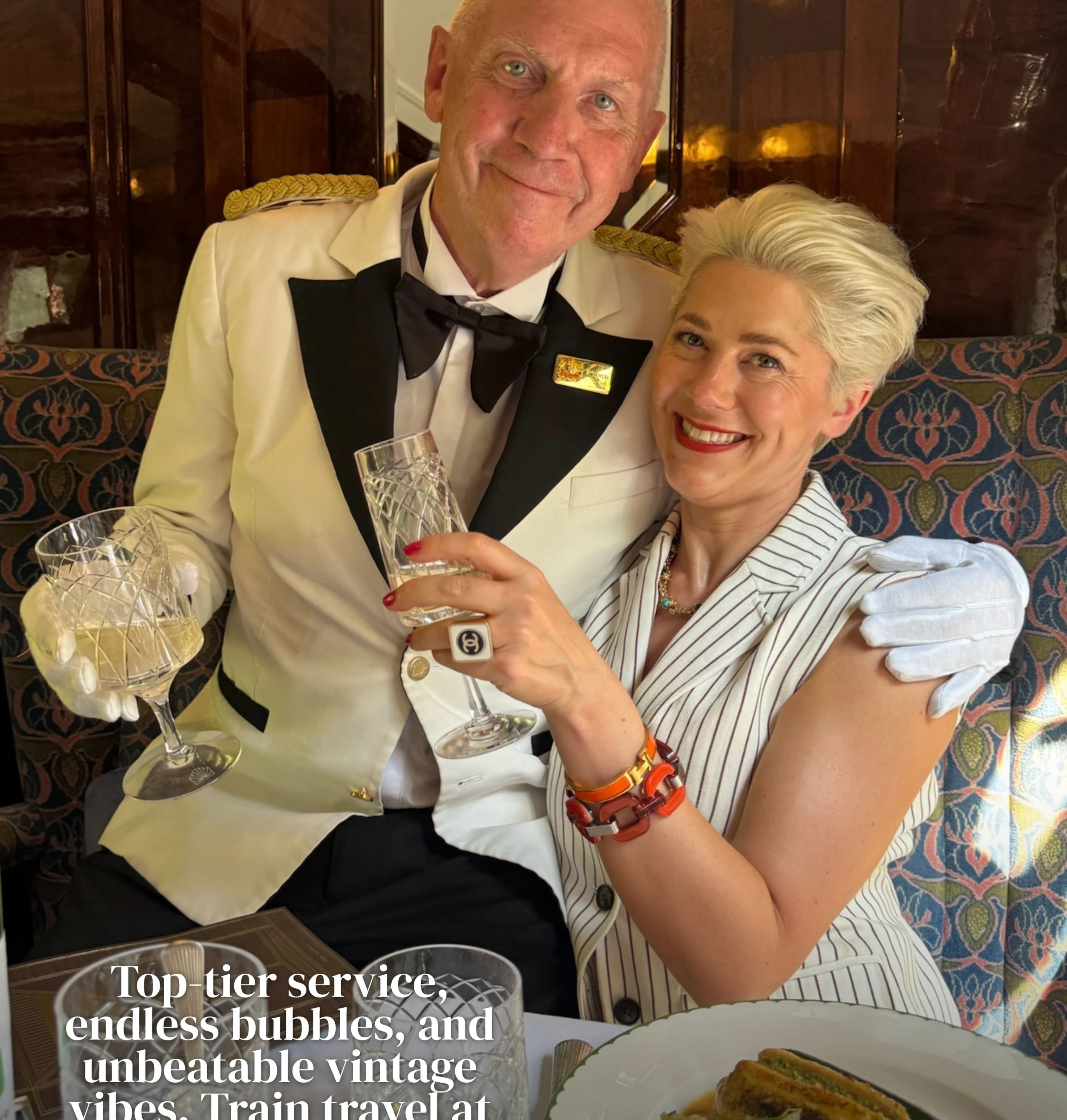 Two people raising crystal glasses aboard a vintage train, with a plated meal on the table
