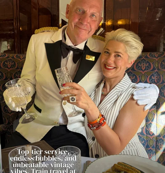 Two people raising crystal glasses aboard a vintage train, with a plated meal on the table
