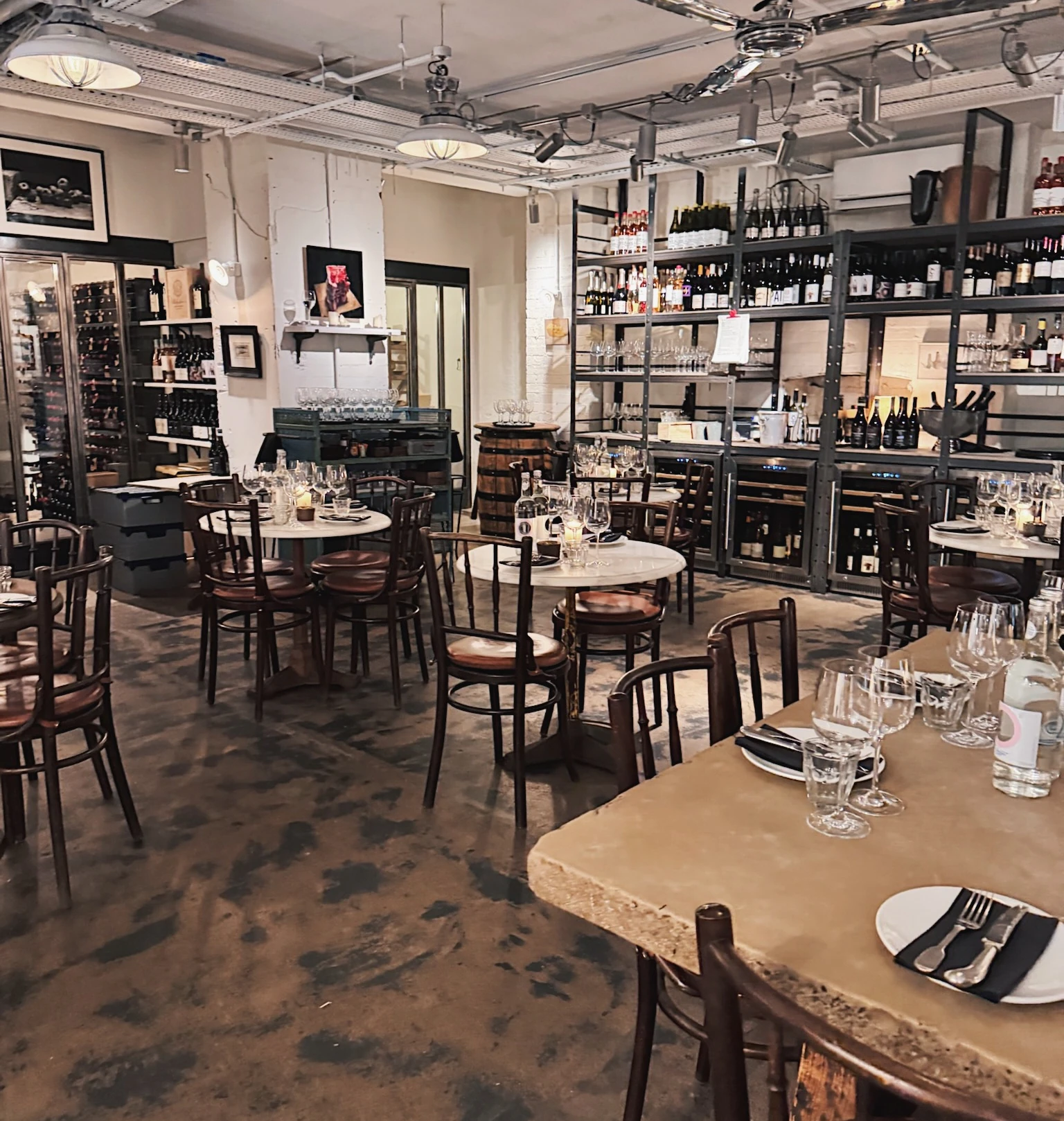 Wine bar interior with stocked shelves, set tables, wine glasses and exposed industrial-style ceiling