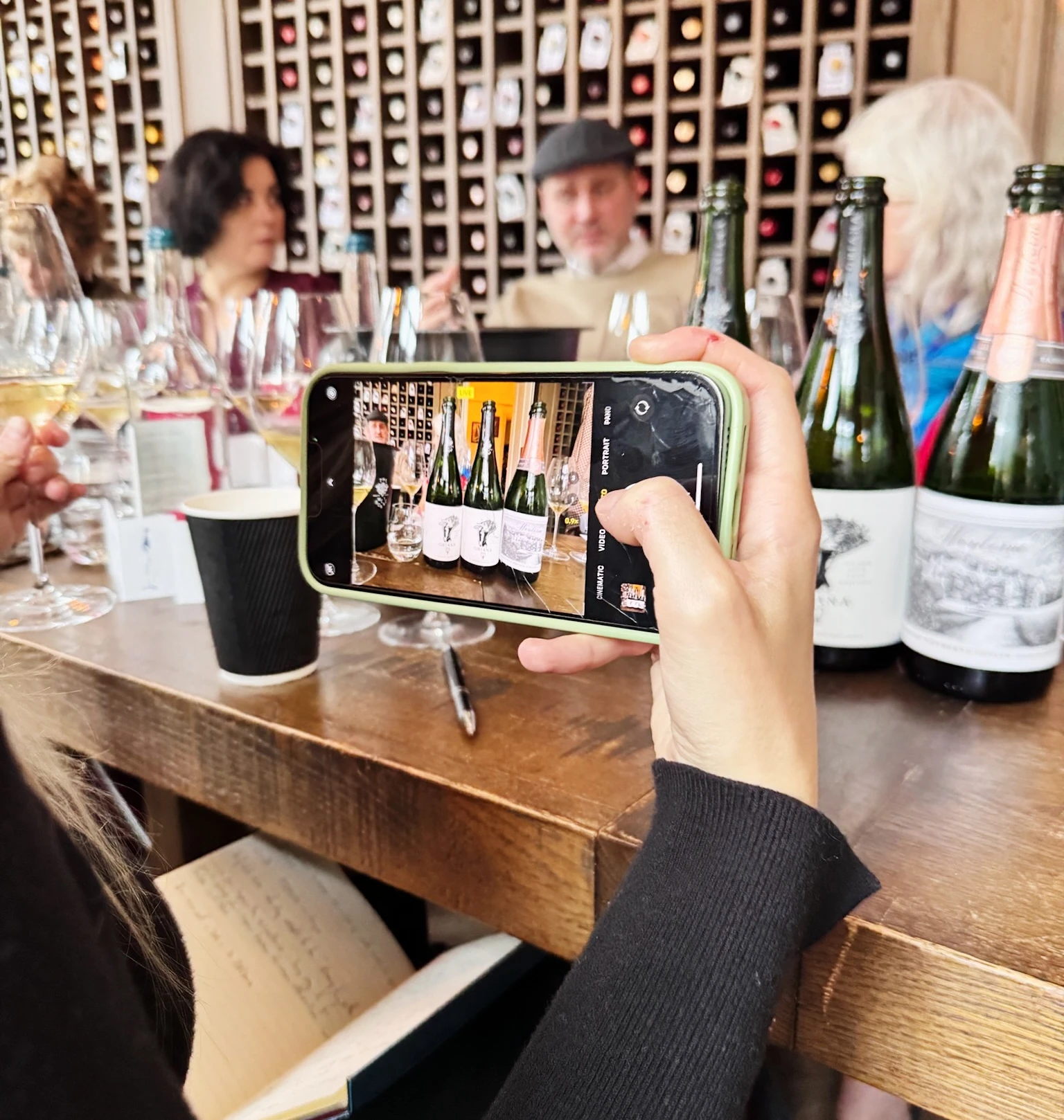 Guests taste sparkling wines beside a wall of wine bottles while one person records the table on a phone