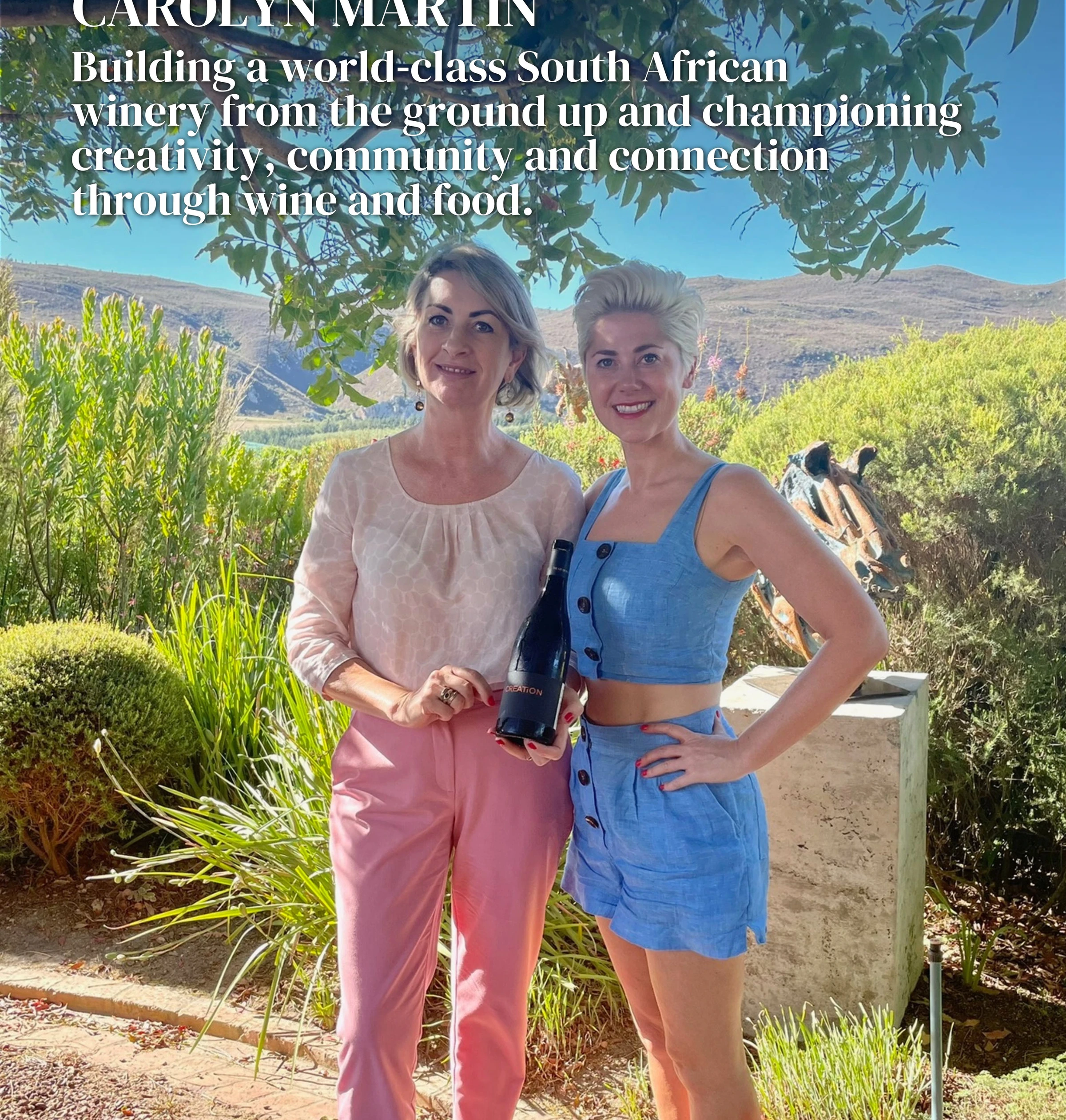 Two women holding a wine bottle in a landscaped South African setting
