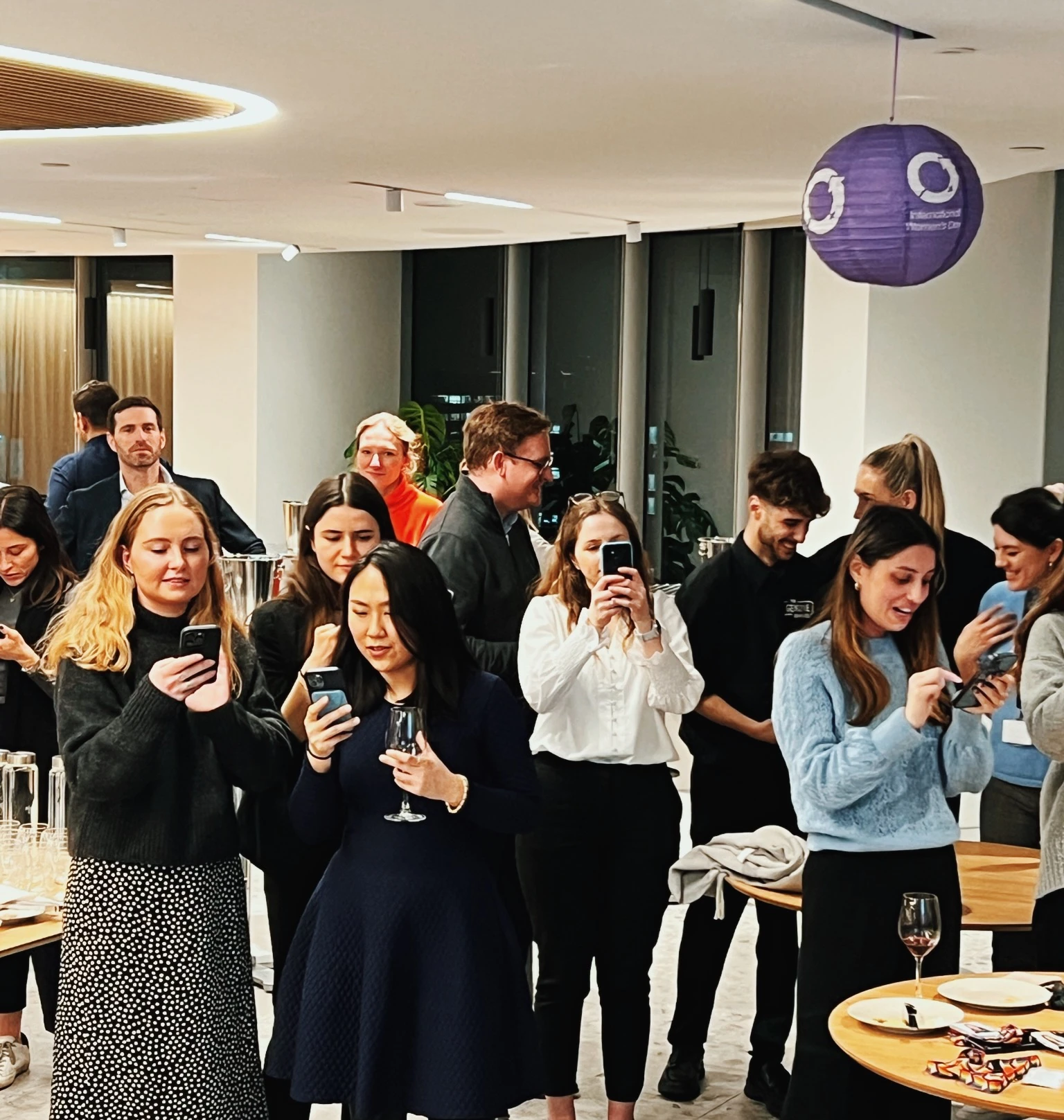 Guests mingle at a wine event, holding glasses and phones beneath a purple hanging decoration