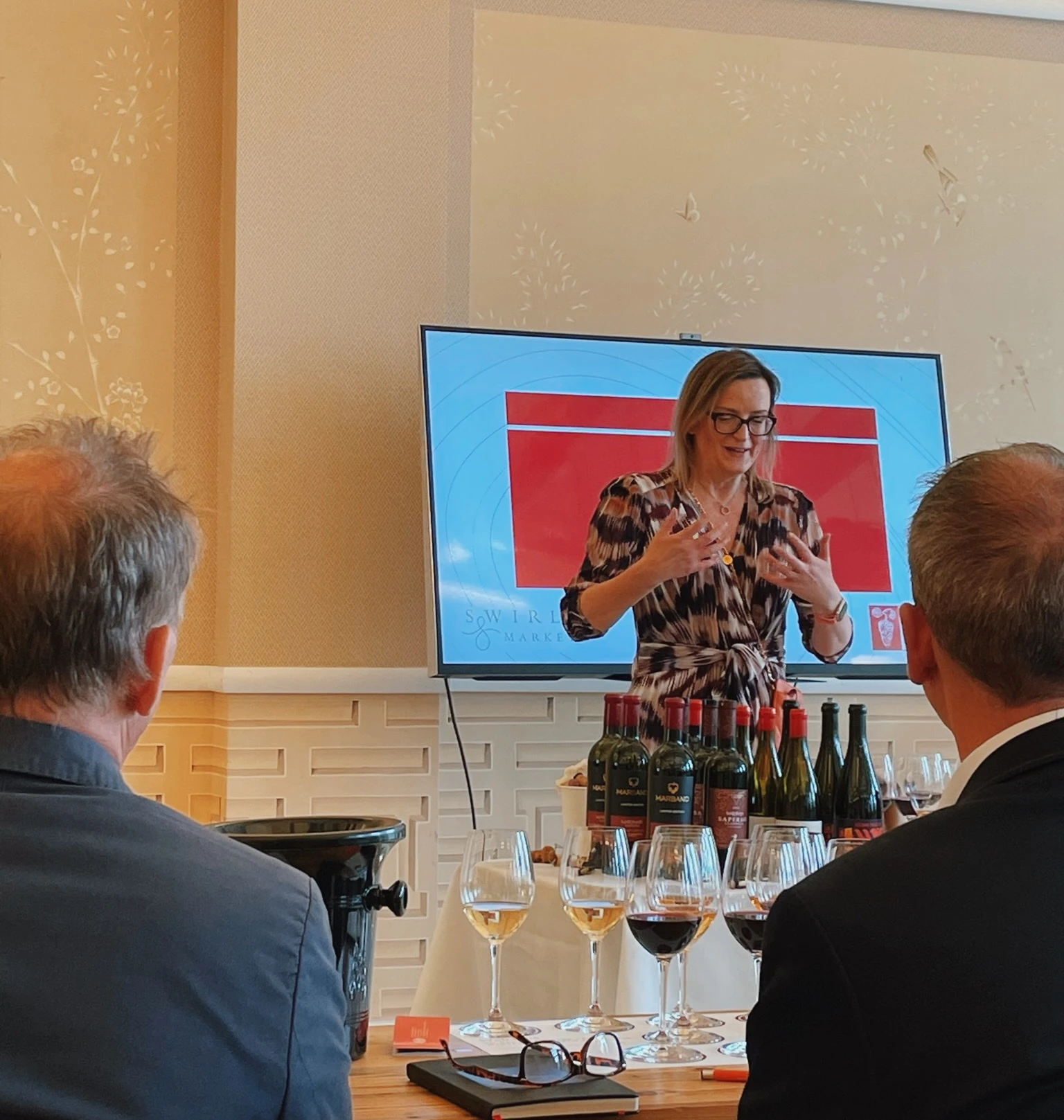 Wine tasting presenter speaking beside bottles and glasses in front of two seated guests