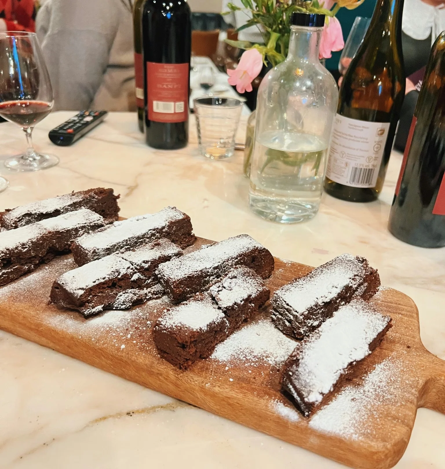 Brownie slices dusted with icing sugar beside wine bottles, glasses and flowers