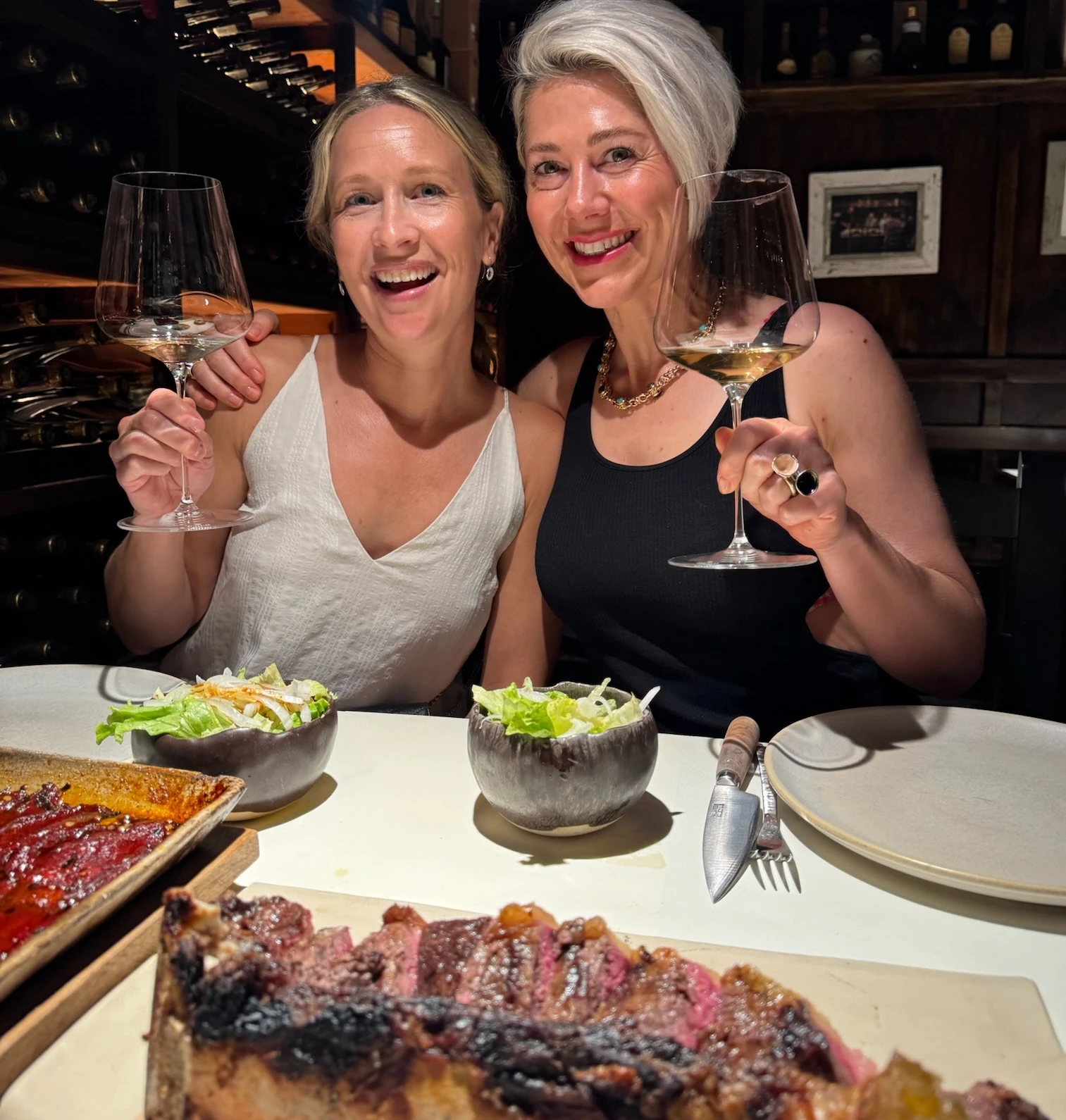 Two women raise wine glasses beside a large grilled steak and salad at Casa Julián Tolosa