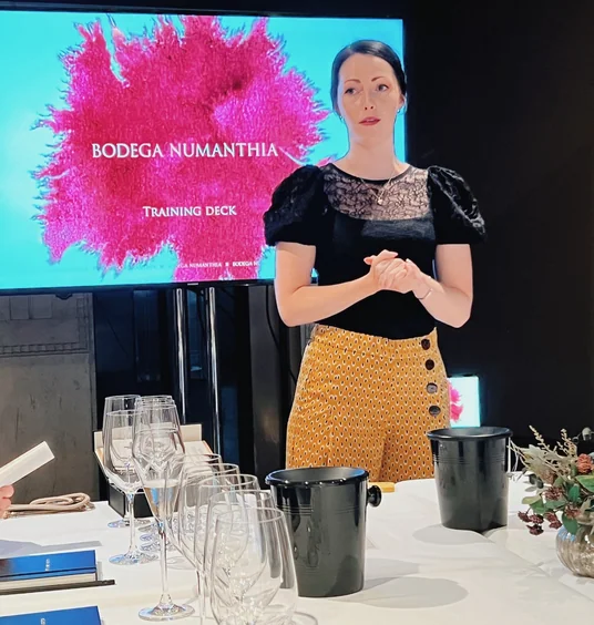Woman presenting beside a screen showing the Bodega Numanthia training deck, with wine glasses arranged on a table