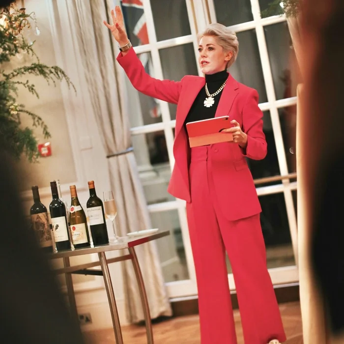 Woman in a pink suit presents at an event beside a table of wine bottles, holding a tablet.