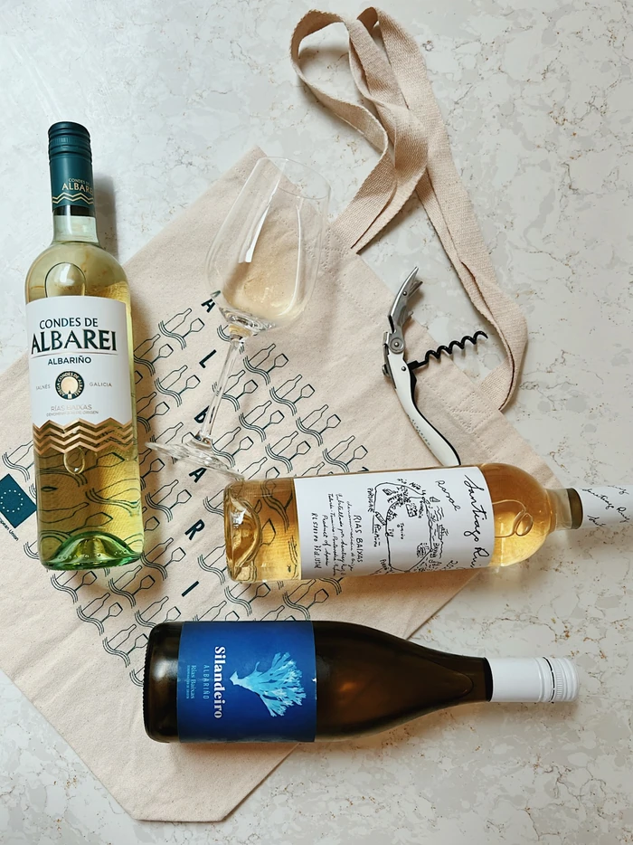 Three Albariño wine bottles, a tasting glass and corkscrew arranged on a patterned cloth