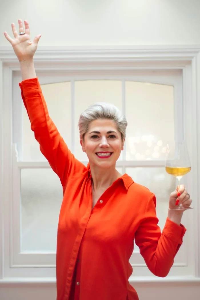 Smiling woman in a bright orange blouse raising one hand while holding a glass of white wine