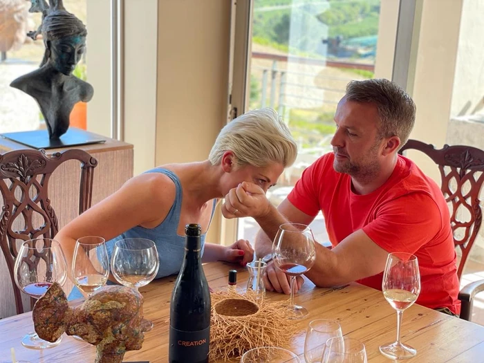 Two people taste wine at a dining table with several glasses and a bottle