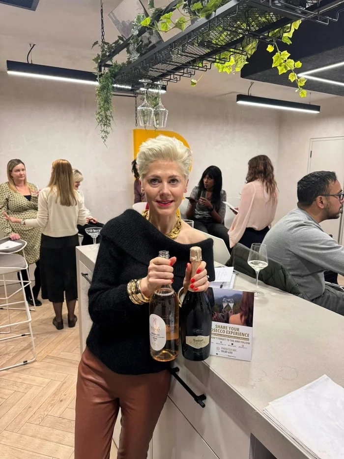 Host holds two Prosecco bottles at a tasting event, with guests and hanging glasses behind her