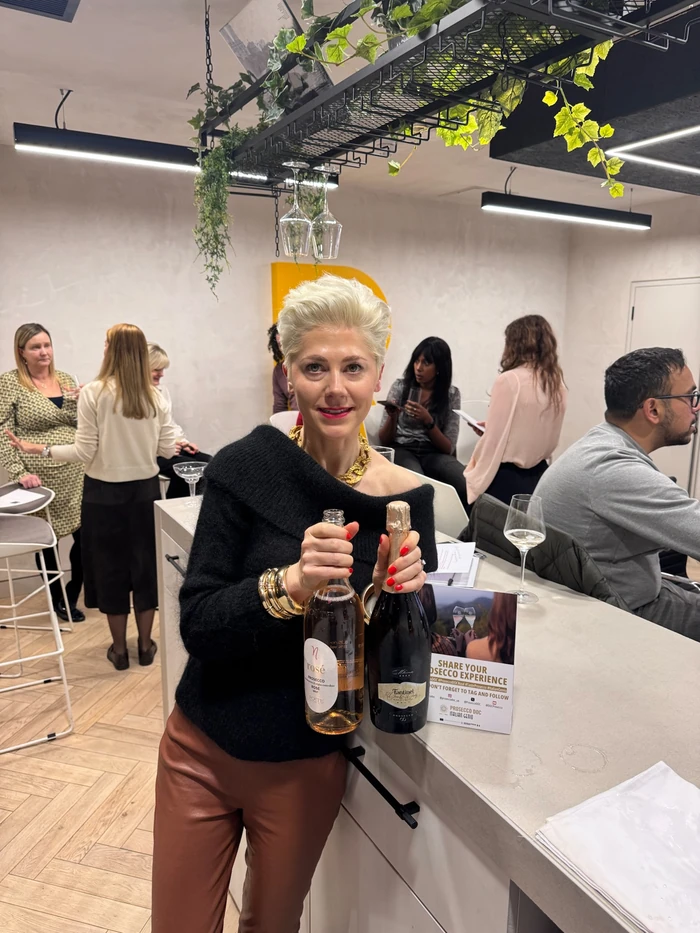 Host holds two Prosecco bottles at a tasting event, with guests and hanging glasses behind her