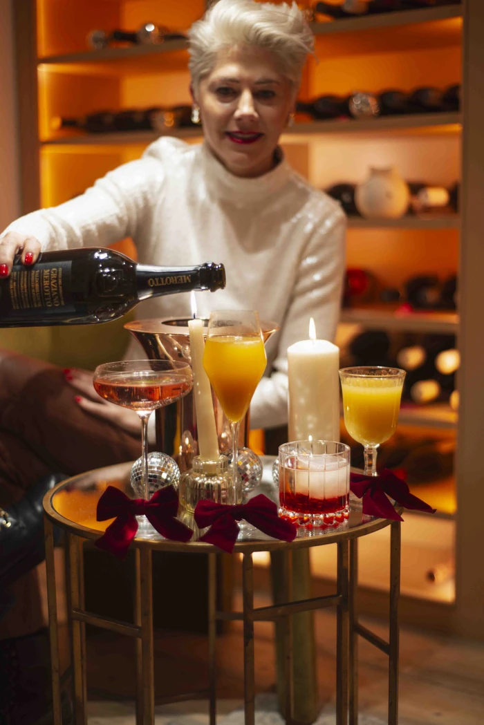 Person pours sparkling wine into a cocktail glass beside Christmas drinks and candles