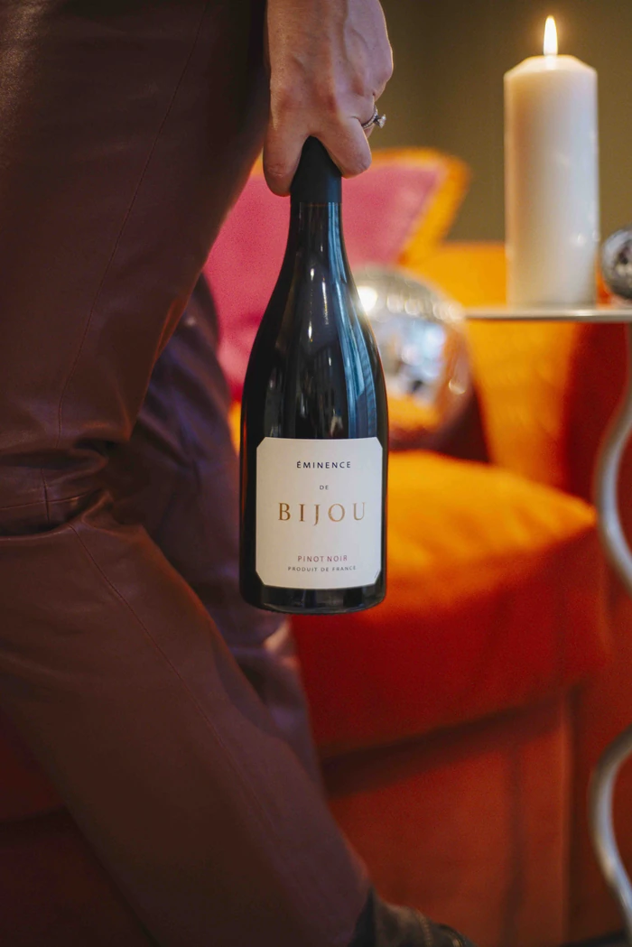 Hand holding a Bijou Pinot Noir bottle beside a lit candle and wine glasses
