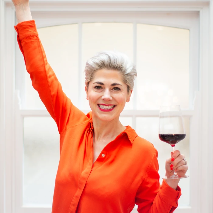 Smiling woman in a bright orange blouse raises one arm while holding a glass of red wine
