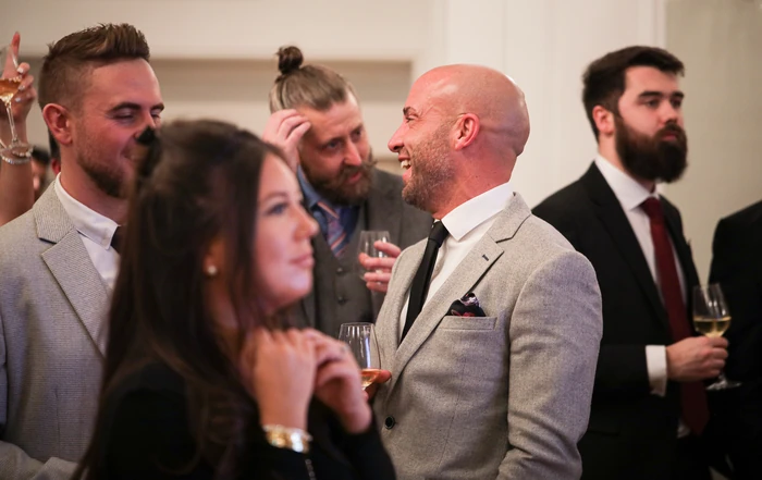 Guests in formal clothing chat and hold wine glasses at an indoor tasting event