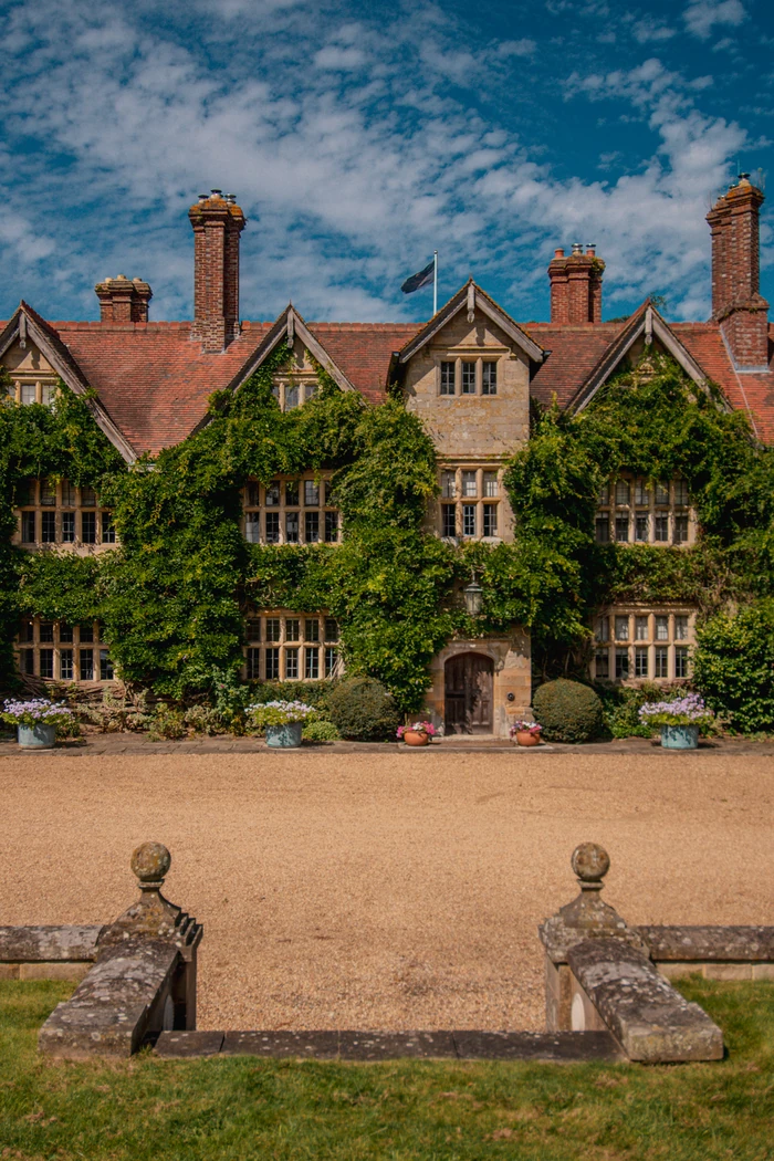 Historic ivy-covered house with ornate chimneys, gardens and a gravel forecourt under a blue sky