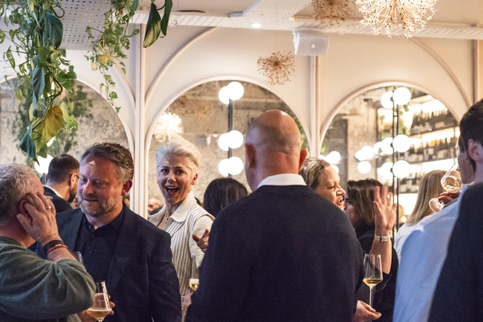 Guests mingle with glasses of wine in a warmly lit venue with arched mirrors and hanging plants