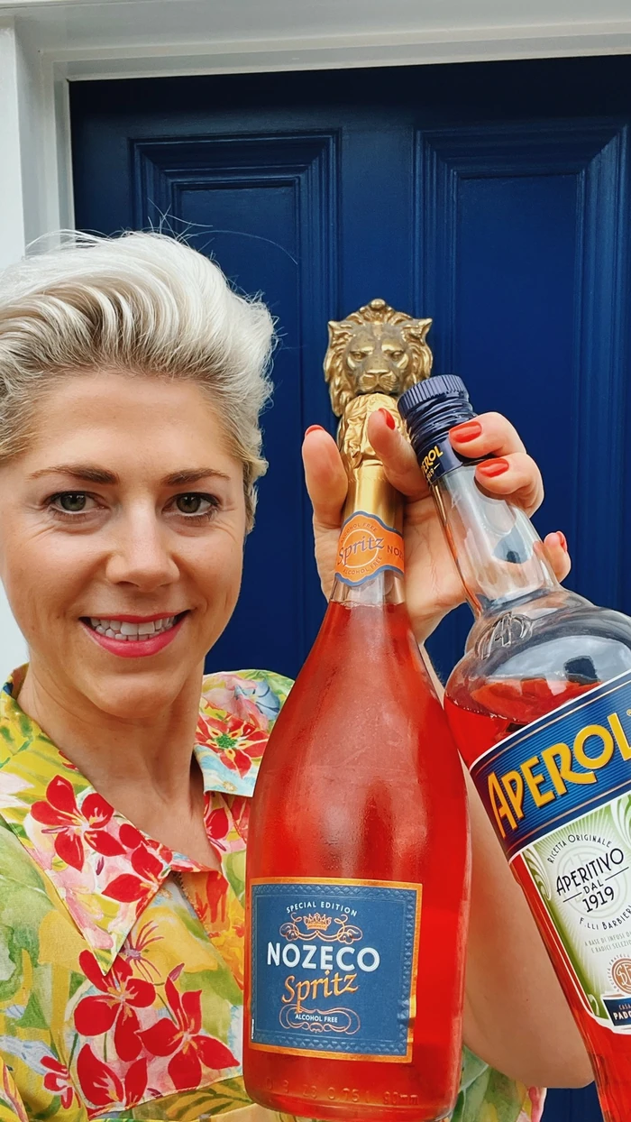 Smiling woman holding Nozeco Spritz and Aperol bottles in front of a blue door