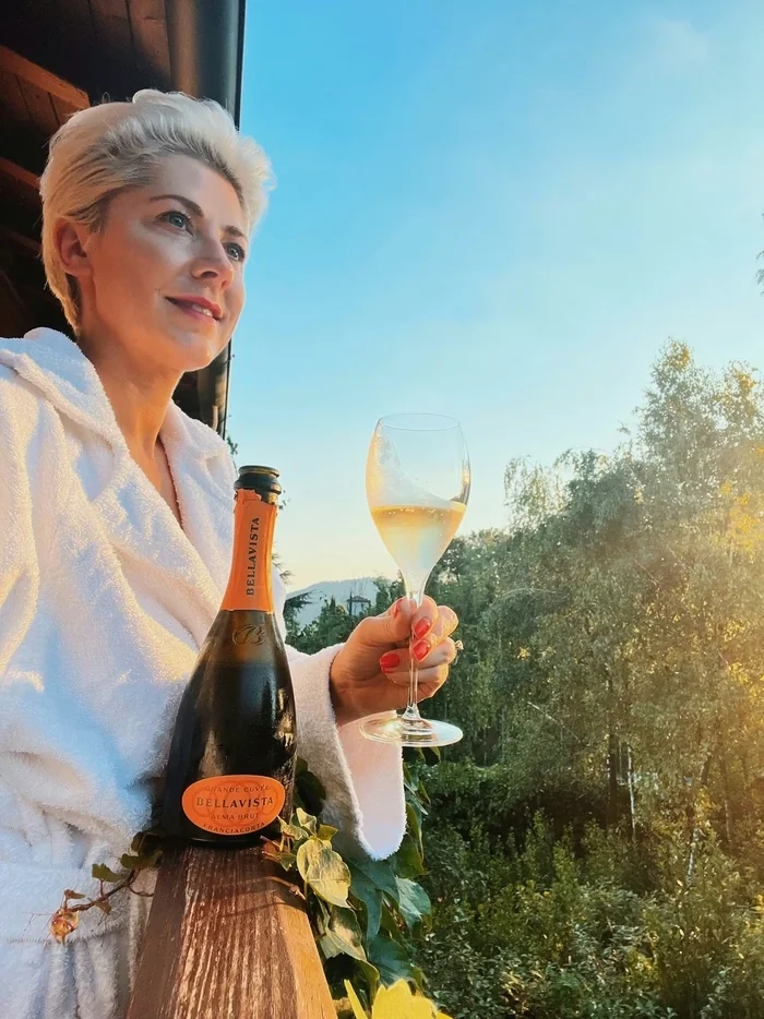Woman in a white robe holding a glass of sparkling wine beside a Bellavista bottle outdoors