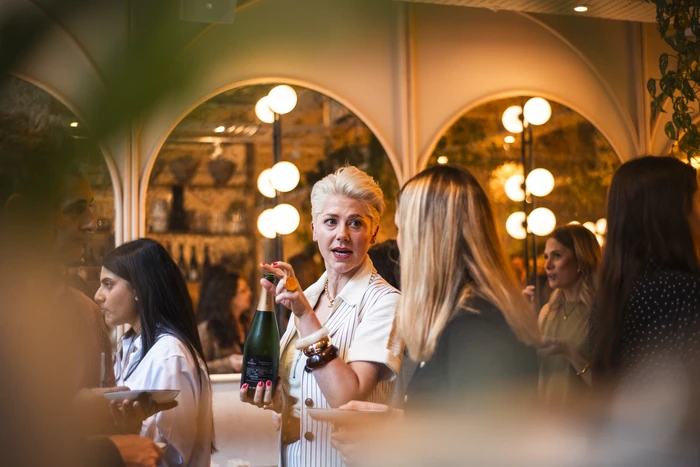 Wine host holding a sparkling wine bottle while speaking to guests at an indoor tasting