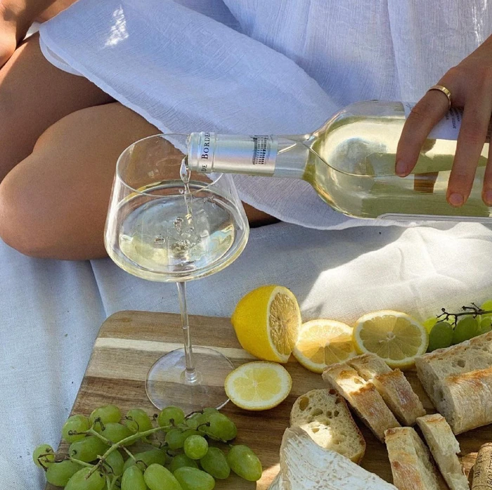 White wine poured into a glass beside lemons, green grapes, bread and soft cheese