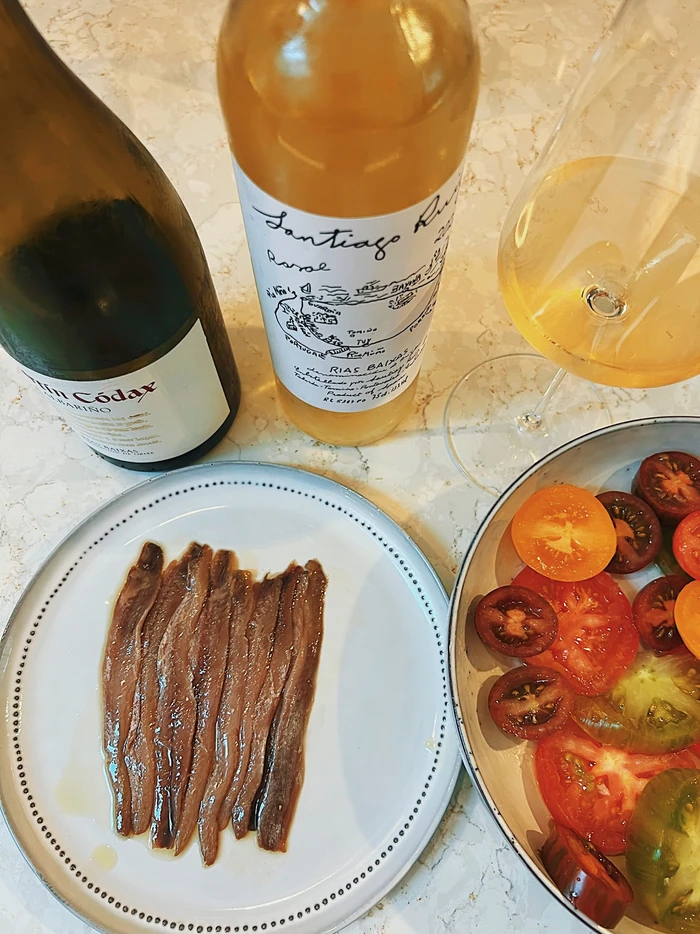 Two bottles of white wine with a filled glass, anchovies and sliced tomatoes on a marble table