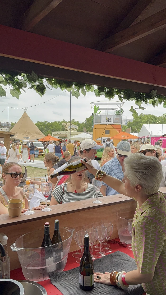 People tasting wine at an outdoor festival bar beneath a leafy canopy