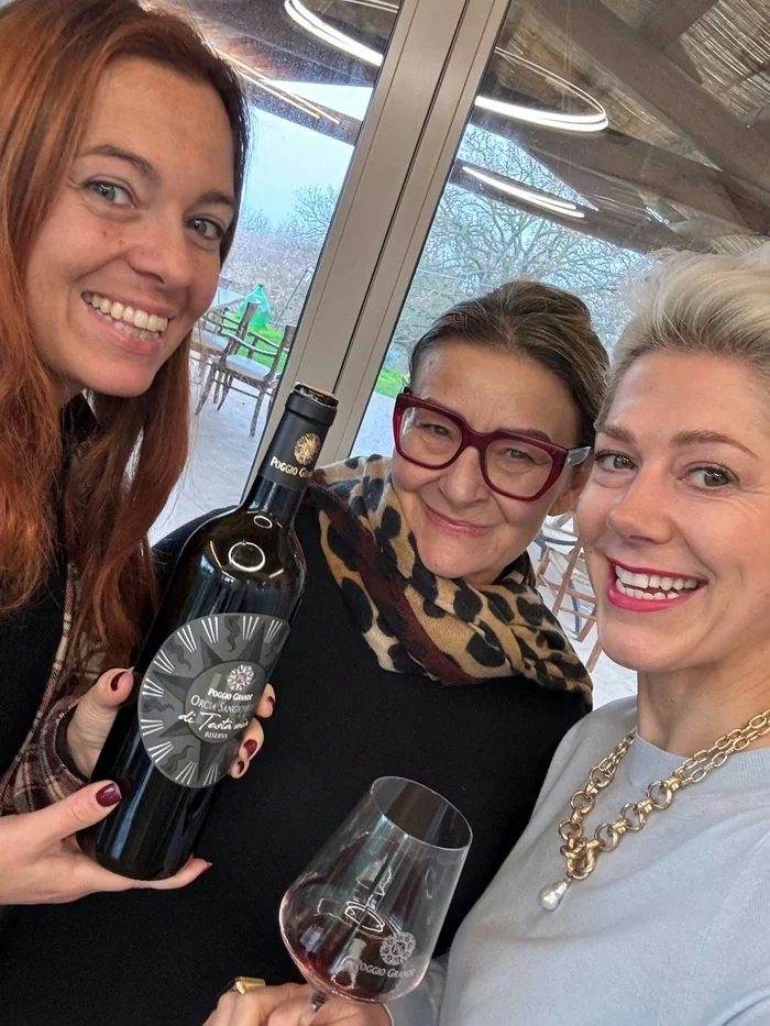 Three women pose indoors with a Poggio Grande wine bottle and a glass of red wine