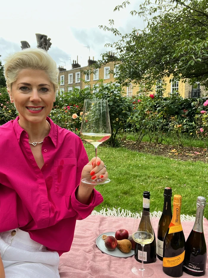 Woman holding a glass of sparkling wine beside bottles and fruit in a rose garden