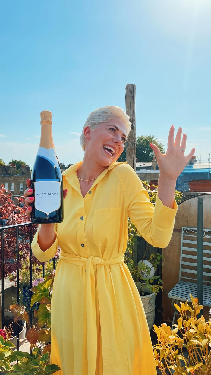 Smiling woman in a yellow dress holding a Nyetimber sparkling wine bottle outdoors