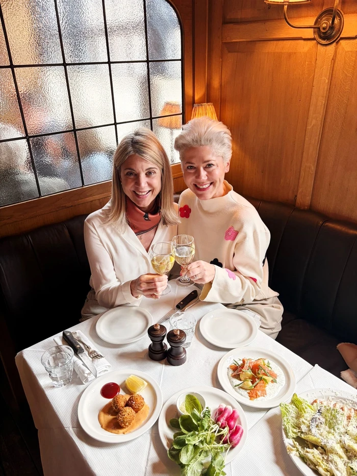 Two women clink glasses of white wine at a restaurant table set with shared dishes