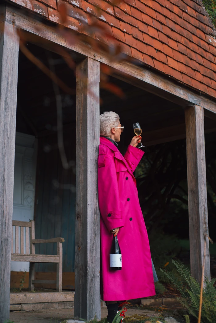 Person in a bright pink coat tasting white wine outside a rustic timber and brick building