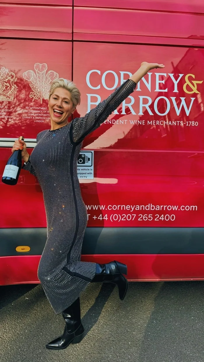 Smiling woman holding a wine bottle beside a red Corney & Barrow vehicle