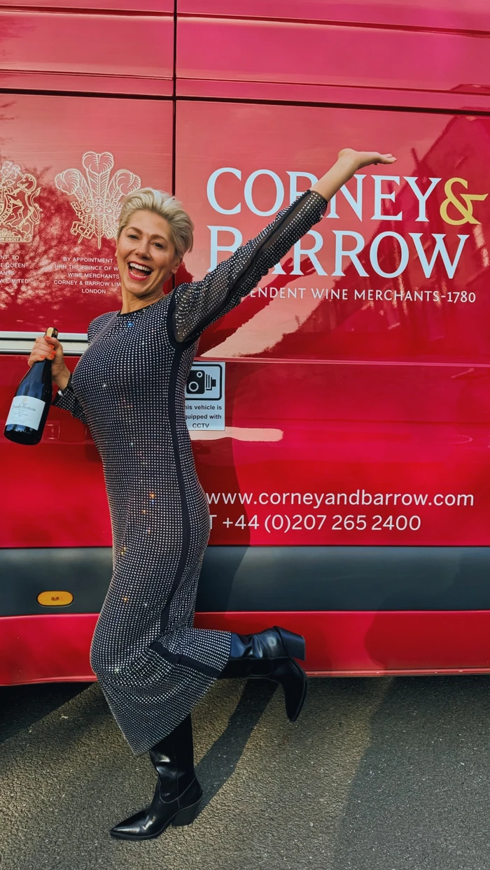 Smiling woman holding a wine bottle beside a red Corney & Barrow vehicle