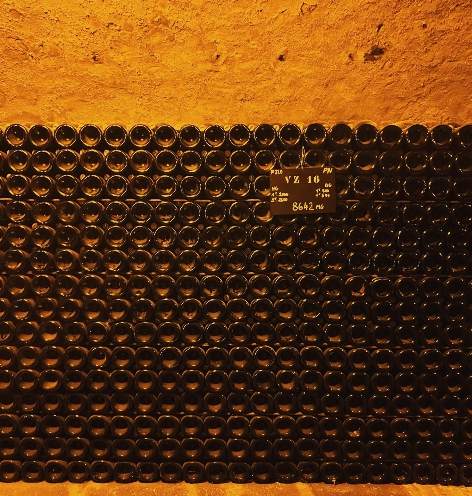 Rows of Champagne bottles stacked in a warm, underground cellar, with a small numbered sign among them.
